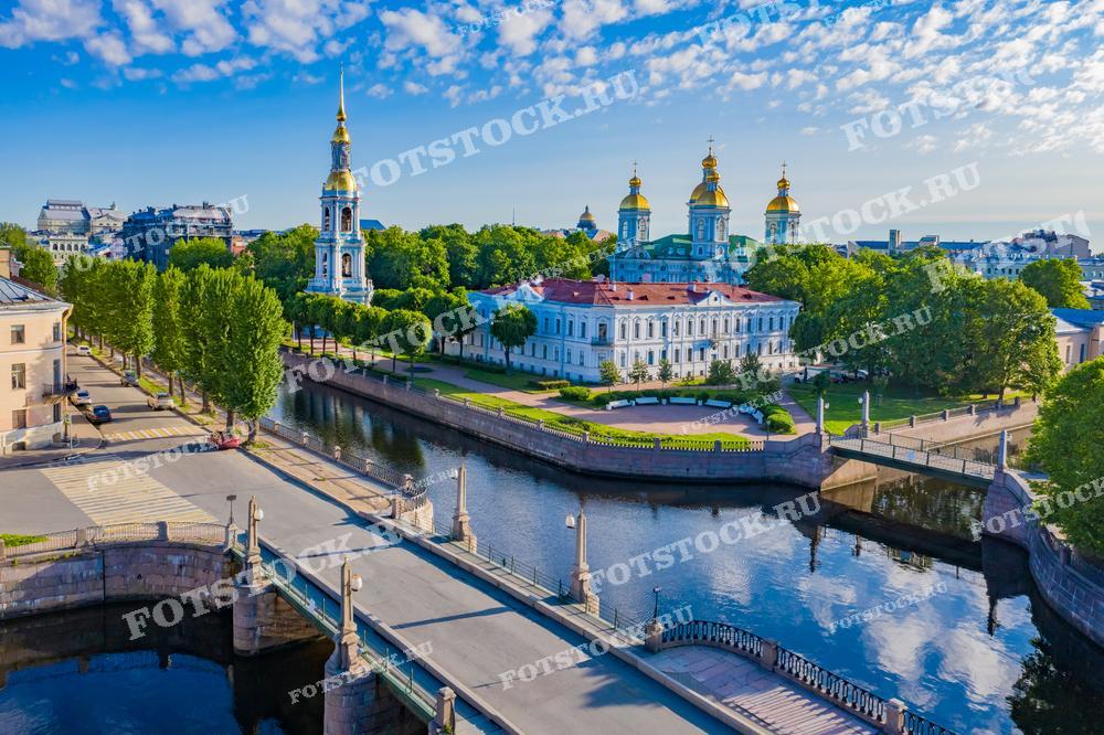 На фотографии изображен Николо-Богоявленский морской собор, окруженный зелеными деревьями, и один из каналов Санкт-Петербурга. Снимок сделан в летний день, что подчеркивается ярким солнцем и голубым небом с облаками. Водная гладь канала отражает архитектуру и зелень, создавая гармоничную картину. Этот вид демонстрирует сочетание природной красоты и исторической архитектуры города.